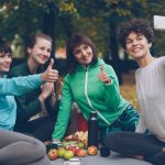 Attractive young ladies yogini are taking selfie using smartphone during picnic in park in autumn. Girls are posing and smiling having fun sitting together on mats.
