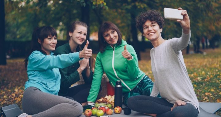 Attractive young ladies yogini are taking selfie using smartphone during picnic in park in autumn. Girls are posing and smiling having fun sitting together on mats.